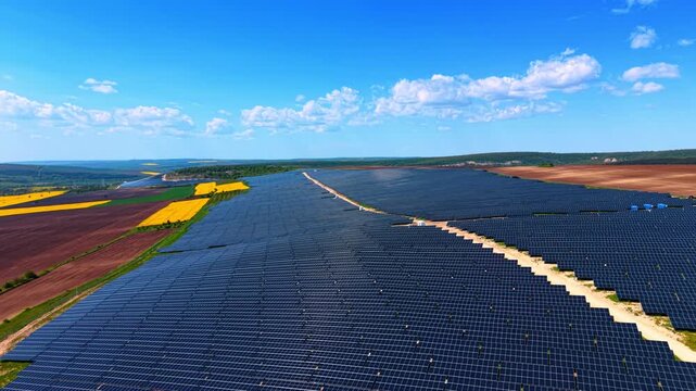 Expansive solar panel rows stretching across rolling farmland under cloudy sky. Wide drone shot of dark blue arrays, rapeseed, and tilled soil with forested hills on the horizon.