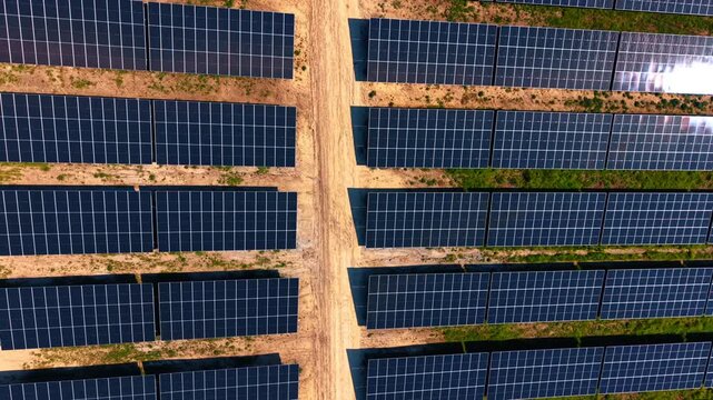 Top down view of solar panel rows separated by dirt pathways. Symmetrical geometric pattern of dark modules, sandy lanes and patches of green grass.