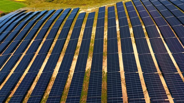 Close aerial view of parallel solar panel rows on dry ground. Low drone angle showing dark modules, sandy soil strips and green hillside edge.