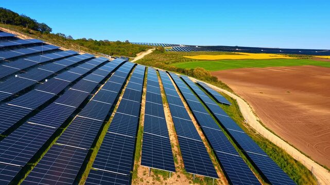 Aerial view of solar farm bordered by plowed and rapeseed fields. Drone perspective showing dark panel rows between brown soil and yellow crops.