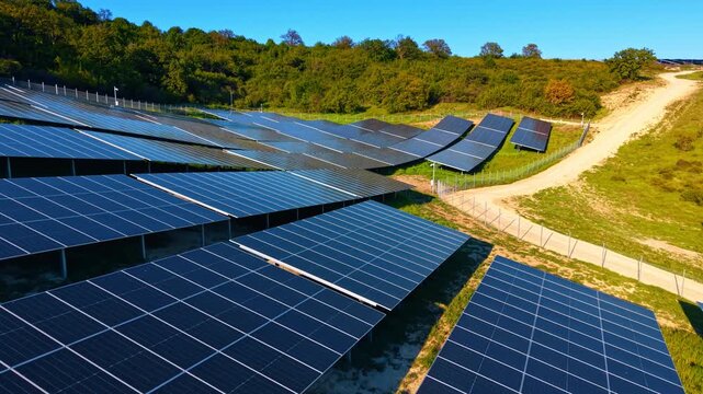 Ground mounted solar arrays near wooded hillside with dirt road. Elevated drone view of fenced panel clusters, green forest and sandy access paths.