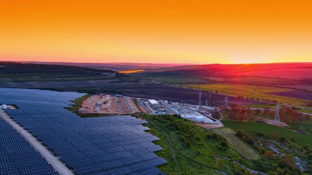 Solar farm and electrical substation under vivid sunset sky. Wide drone view of panel rows, pylons, construction site, and green rolling hills.