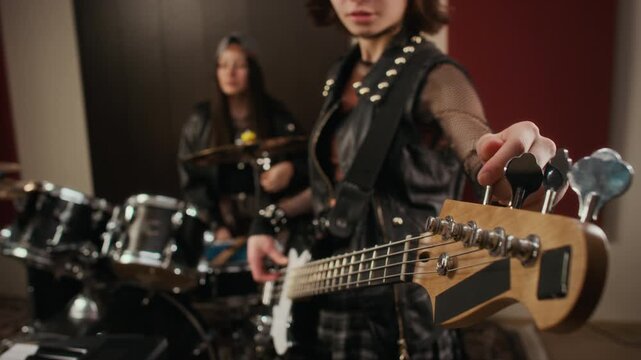 Caucasian teenager tuning bass guitar during band rehearsal in music studio with drummer in background. Footage is perfect for music education promos and youth lifestyle commercials