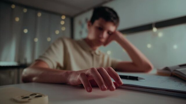 A tired and bored teenage boy taps his fingers on the table, feeling unmotivated while doing his homework late at night in his room.