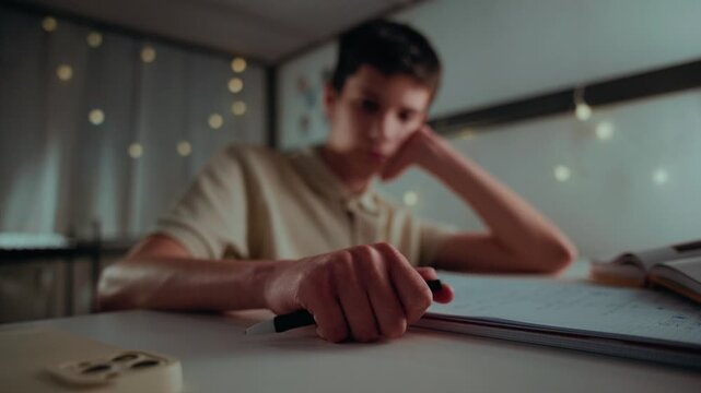 Close up of a bored student's hand tapping a pen on a notebook while studying late at night. He appears tired and unmotivated, struggling with his homework.