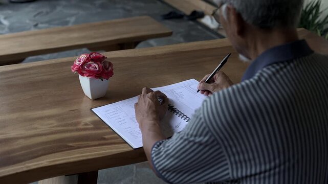 Over shoulder back shot of Asian old man writing a book on wooden table