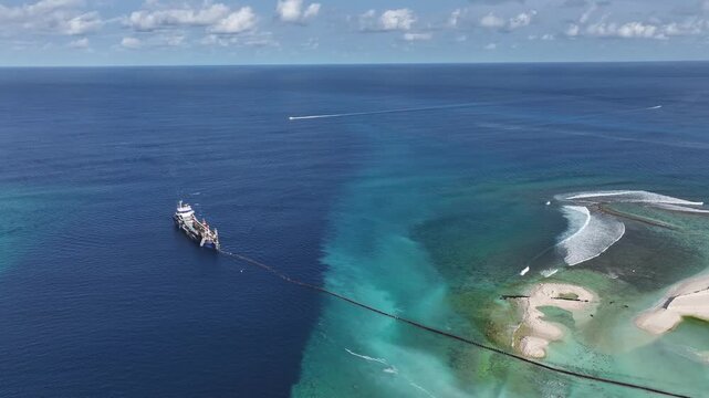 Aerial view of an industrial dredging vessel operating in the turquoise waters near a coral reef under a blue sky with scattered clouds in Gulhi, Kaafu Atoll, Maldives.