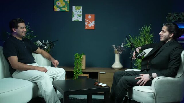 Two men converse and laugh in a modern studio lounge setup with microphones