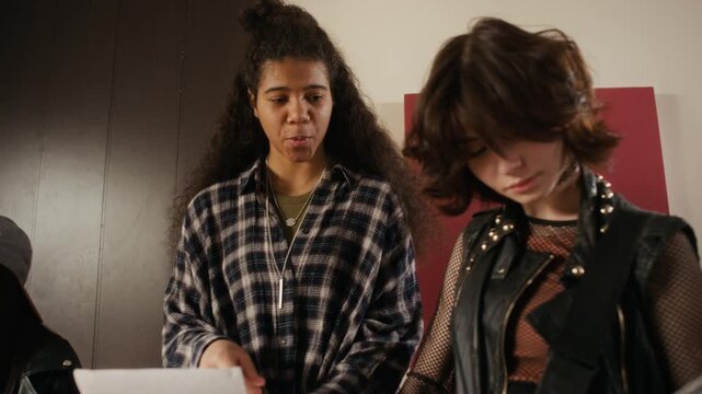 Multiethnic female teenagers rehearsing rock music in studio, playing drums and bass guitar while reading lyrics. Video is suitable for youth culture, music education, and diversity campaigns