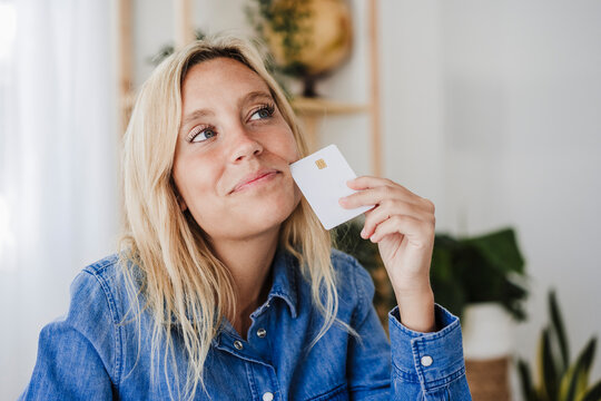 Thoughtful blond woman holding credit card at home