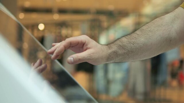 Hand taps glass screen in modern store. Focused finger moves across glossy surface. Background blurred, warm lighting. Ideal for retail, tech, or shopping ads. Shows interaction with digital displays