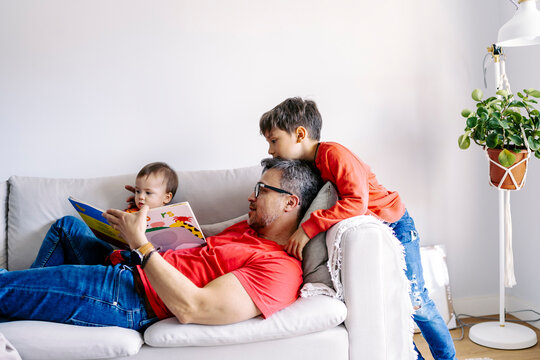 Father reading book with sons on sofa in living room at home