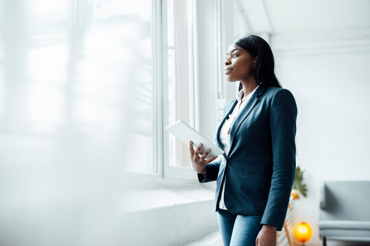 Thoughtful businesswoman holding tablet PC looking through window in office
