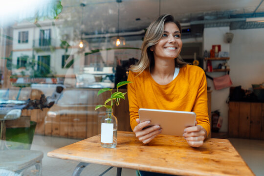 Happy woman sitting with tablet computer in cafe seen through glass