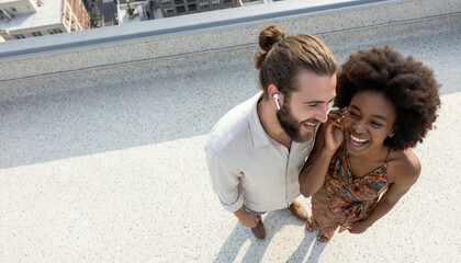 Fototapeta premium High angle view of a happy young couple standing on a rooftop, laughing and sharing wireless earphones while listening to music together on a sunny day