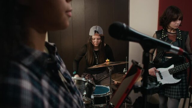 African American female teenager singing into microphone with Caucasian teen bandmates playing drums and bass in studio. Perfect for youth culture and music education marketing campaigns