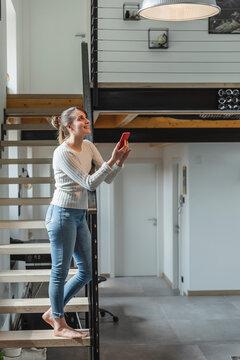 Smiling woman controlling home functions with smart phone while standing on steps at home