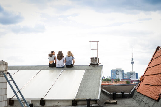 Germany, Berlin, back view of three friends sitting side by side on roof top