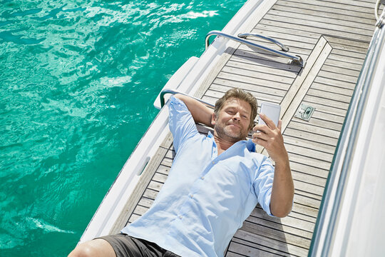 Smiling man lying on deck of his motor yacht checking emails