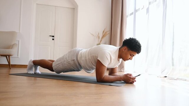Young man exercises in plank position on a yoga mat in a well-lit room, with a smartphone beside him for workout tracking