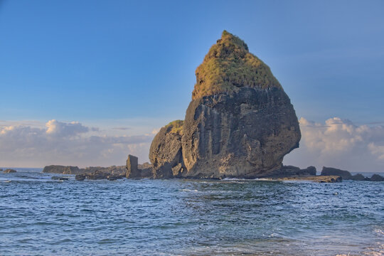 Towering volcanic sea stack rock with a pointed peak rising dramatically from ocean waters under a clear blue sky. Papuma Beach, Jember, Indonesia. 