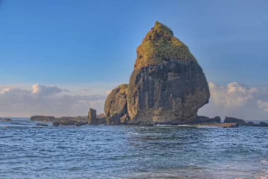 Towering volcanic sea stack rock with a pointed peak rising dramatically from ocean waters under a clear blue sky. Papuma Beach, Jember, Indonesia.