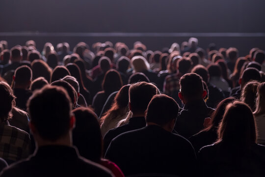 Large seated audience watching performance in dim auditorium, silhouettes and backlit heads create quiet, focused atmosphere.