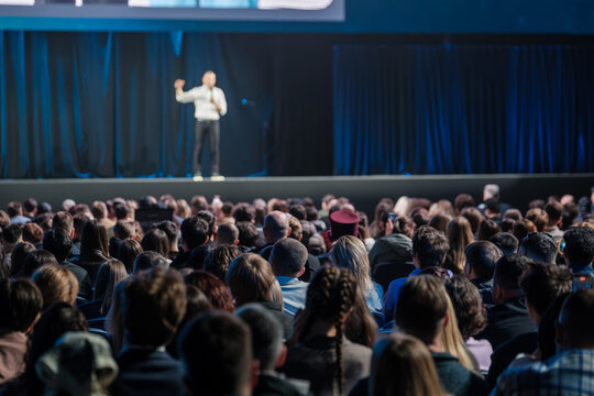 Large crowd listening to keynote speaker on stage at professional conference, moody blue lighting, focused atmosphere.