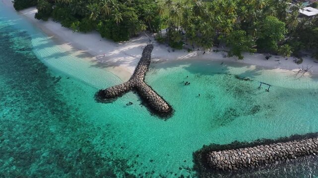 Aerial view of a tropical beach with a stone breakwater, turquoise water and palm trees in Kendhoo, Baa Atoll, Maldives.