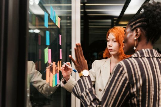 Two colleagues brainstorm at a glass wall in a modern office, arranging colorful sticky notes and planning ideas during a collaborative team session.