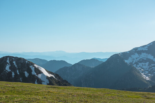 Scenic view from sunlit grassy meadow to abyss of alpine valley between big rocky mountains with snows and hilly layers of ridge silhouettes far away under clear blue sky in bright sun in sunny day.