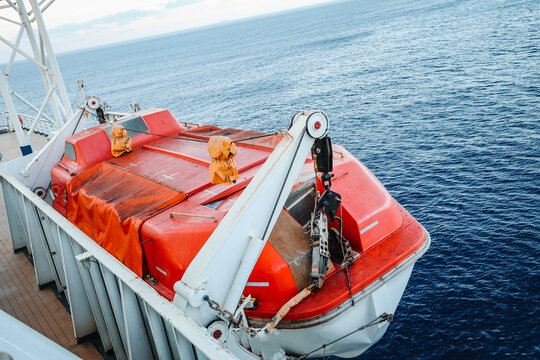 High angle view of an orange enclosed emergency lifeboat marked for maximum 150 persons, mounted on a passenger vessel deck with the blue sea in the background.