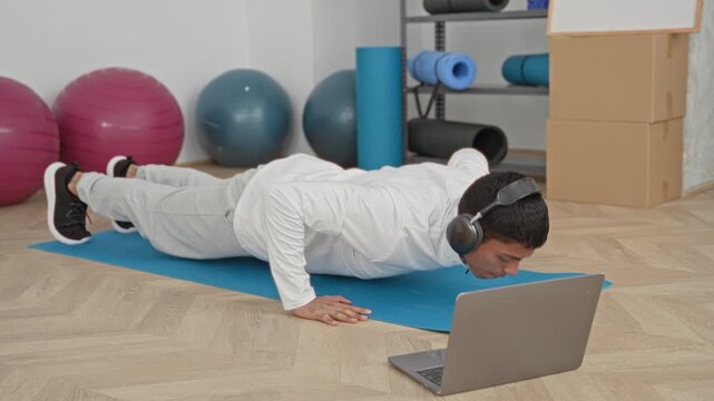 Young hispanic man doing pushup on blue mat with laptop and headphones in studio; home workout determination focus.