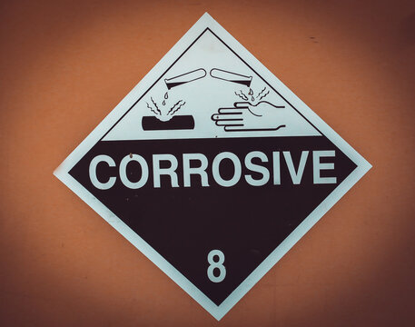 Diamond-shaped Class 8 Corrosive hazard warning placard with test tubes, hand, and surface burn pictograms mounted on a metal wall.