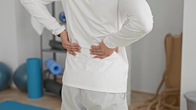 Young hispanic man in white top holding hands on lower back for backache in yoga studio with mat and foam roller visible during training session; discomfort recovery.