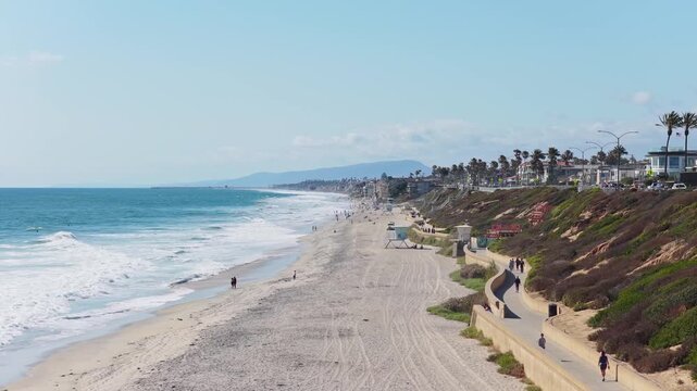 Scenic aerial of Carlsbad State Beach in California showing pedestrians walking on seaside promenade and Pacific Ocean waves crashing on sandy shore