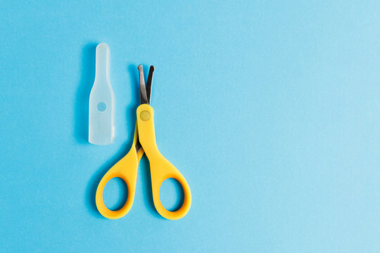 Yellow baby safety scissors with rounded tips and plastic protective cover cap on bright blue background, horizontal flat lay shot of newborn nail care trimmer tool with large copy space