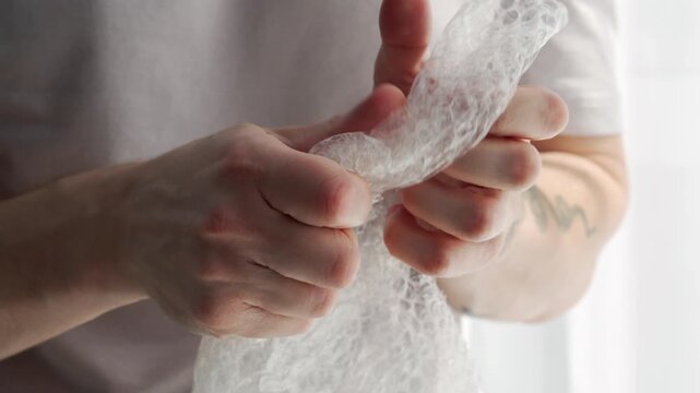 A guy holds bubble wrap.
He pops small bubbles on the transparent film with his fingers.