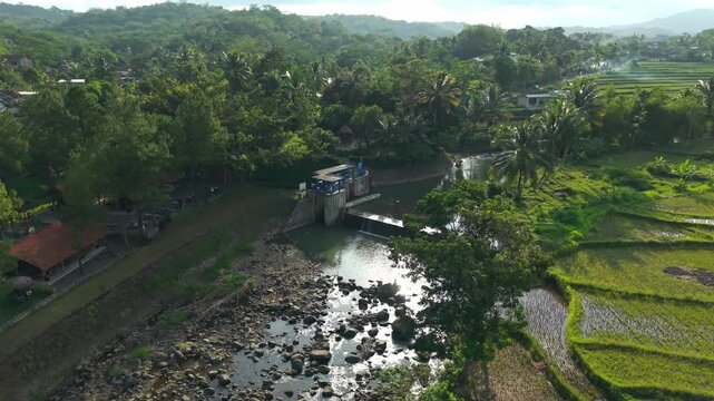 Aerial view of Tangsi River Water Gate surrounded by lush tropical trees and terraced rice paddies under bright sunlight in Kecamatan Salaman, Jawa Tengah, Indonesia.