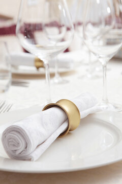 Setting festive table with white tablecloth. White serving napkin with yellow metal ring is placed on plate. Empty glasses for drinks. Concept of festive events. Preparation for the reception of