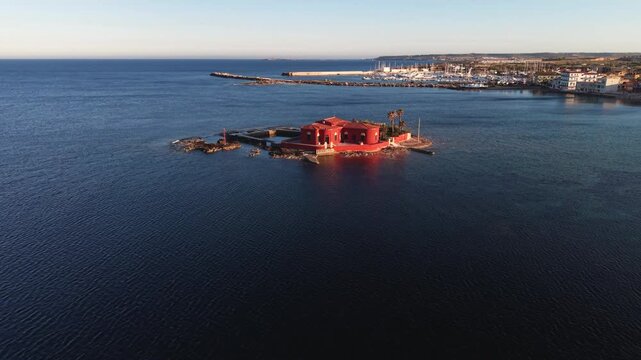 Aerial view of the red villa on Isolotto Brancati surrounded by the blue Mediterranean Sea during golden hour in Marzamemi, Sicilia, Italy.