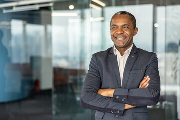 Obraz premium Confident professional businessman standing with folded arms in a modern corporate office, looking away and smiling, representing leadership, success, and executive excellence