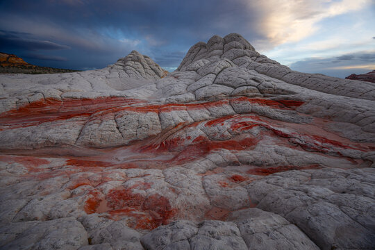 View of the unique White Pocket sandstone formations with red swirls and brain-like textures under a dramatic sunset sky in Coconino County, Arizona, United States.