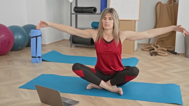 Woman seated crosslegged on mat with laptop, bare feet visible, stretching arms in a yoga studio; serenity focus wellness.
