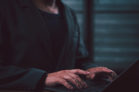Unrecognizable professional person wearing blazer typing laptop keyboard inside dim office space late night performs corporate work tasks focused digital management duties alone online across wooden.
