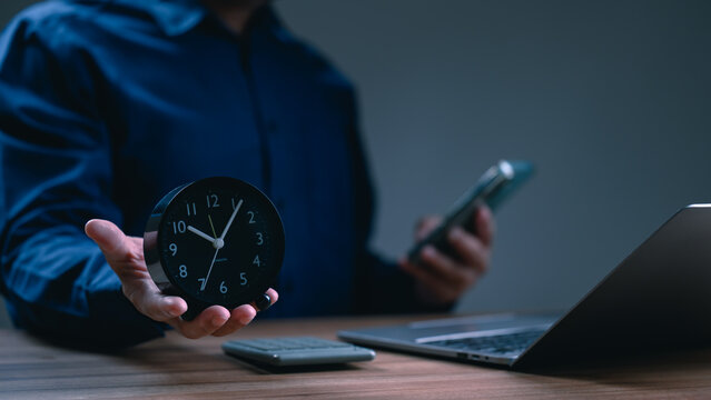 Businessman holding black alarm clock smartphone sitting wooden desk near laptop calculator inside dark office room managing time schedule deadlines overtime productivity late night evening work.