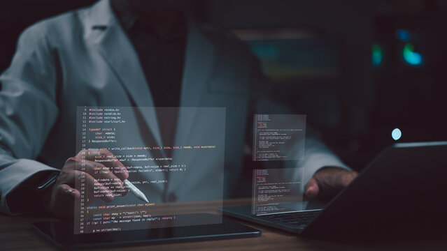 Professional software developer wearing gray suit jacket operates stylus pen tablet laptop computer digital programming code overlays appearing dark office environment late night work session screen.