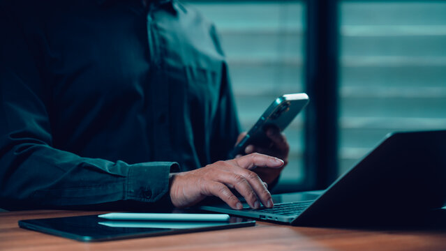Professional man wearing dark shirt types laptop keyboard holding smartphone beside tablet wooden desk inside dim office space late night performing digital multitasking work task corporate project.
