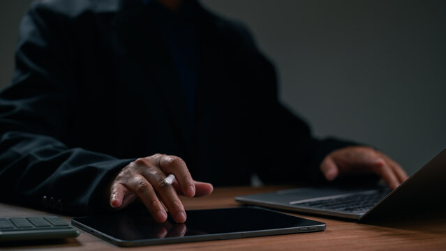 Professional businessman wearing dark suit jacket operates digital tablet stylus pen beside laptop computer sitting wooden desk inside dim office environment night work session period research.