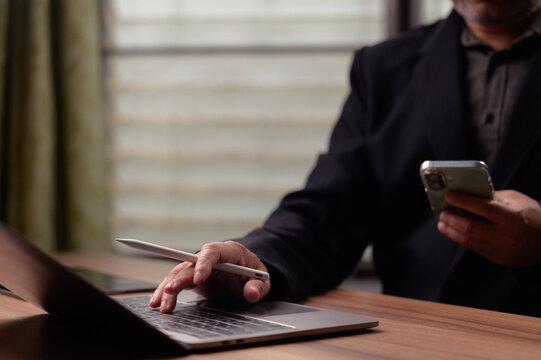 Professional businessman wearing black suit jacket types laptop keyboard when holding smartphone stylus pen wooden desk indoor office space multitasking digital nomad remote work executive managing.
